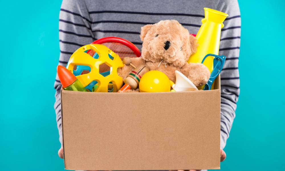Male volunteer holding a donation box full of toys, including a ball, a plush bear, sunglasses, and other small games.